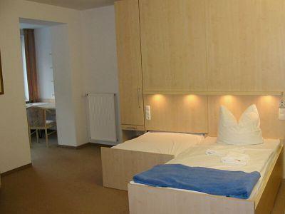 Ein minimalistisch gestaltetes Schlafzimmer mit maßgefertigten Möbeln. Das Zimmer enthält ein Einzelbett mit weißer Bettwäsche, über dem kleine Wandleuchten angebracht sind. Daneben befindet sich ein funktionaler Arbeitspla