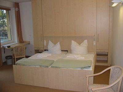 Ein funktional gestaltetes Doppelzimmer mit zwei zusammenstehenden Einzelbetten, ausgestattet mit weißer Bettwäsche und grünen Bettdecken. Die Möbel sind maßgefertigt, darunter ein großer Schrank und ein Arbeitsplatz mit T